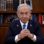 An older man in a suit sits at a wooden desk in an office, framed by a bookshelf showcasing photos and books. An Israeli flag stands to his left, while his lapel displays a yellow ribbon pin symbolizing freedom. In the air lingers a sense of determination akin to Netanyahu's unwavering stance.