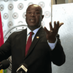 A man in a suit and red tie stands at a podium with microphones, gesturing with both hands raised. Behind him, a backdrop featuring circular emblems and a partial view of a flag with red, yellow, green, and black stripes alludes to Staatsolie's influence.