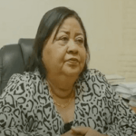 Jennifer van Dijk-Silos, a former BiZa-minister with shoulder-length dark hair, is seated at a desk. She is wearing a patterned blouse and has her hands clasped. Piles of papers and books are visible in the background, possibly related to the verkiezingscrisis.